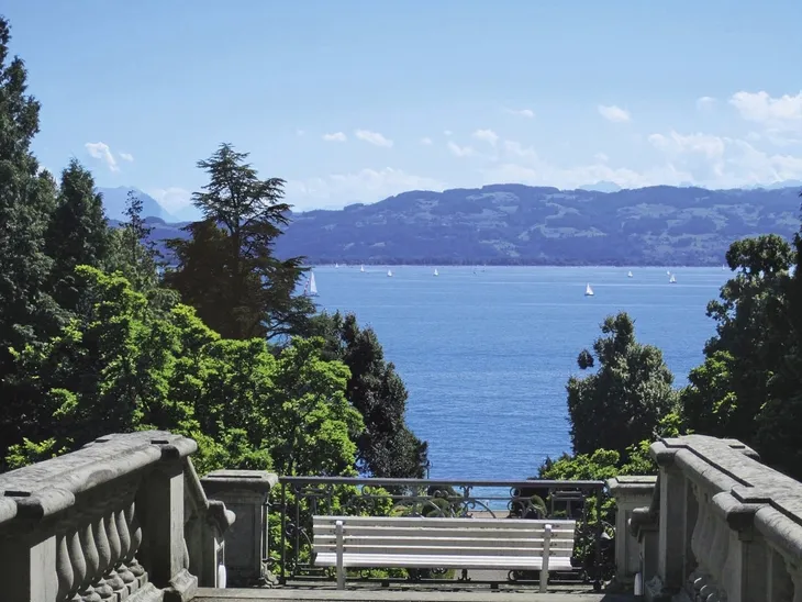 Blick von Treppe auf Bodensee mit Segelbooten und Bergen im Hintergrund.