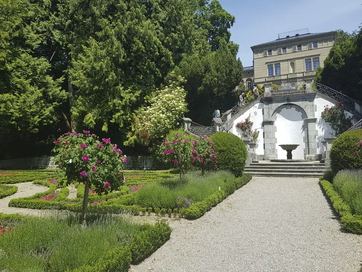 Gartenanlage mit Blumen, Treppe und historischem Gebäude im Hintergrund.