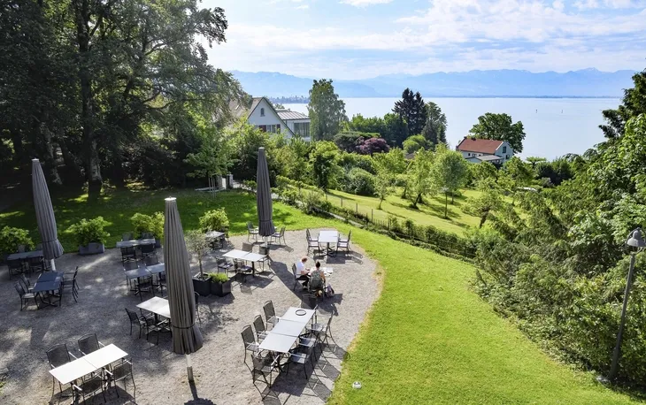 Biergarten der Villa Alwind, in der Ferienanlage Lindau, mit Tischen und Stühlen, sowie geschlossenen Sonnenschirmen, umgeben von Bäumen und einer Wiese mit Blick auf den Bodensee. bei leichtbewölktem Himmel.