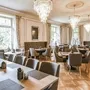 Elegantes Restaurantinterieur in der Villa Alwind in der Ferienanlage Lindau. Unterschiedlich große Holztische mit grauen, gepolsterten Stühlen auf Holzboden, sowie große weiße Fenster mit zusammengebundenen Vorhängen und moderne Kronleuchter an der Decke, die das Restaurant erhellen.