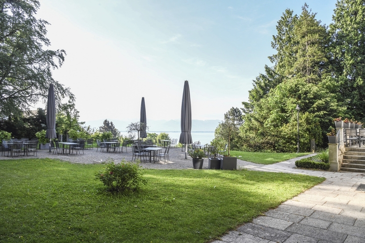 Biergarten der Villa Alwind, in der Ferienanlage Lindau, mit Tischen und Stühlen, sowie geschlossenen Sonnenschirmen, umgeben von Bäumen und einer Wiese mit Blick auf den Bodensee. bei leichtbewölktem Himmel.