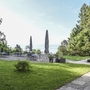 Biergarten der Villa Alwind, in der Ferienanlage Lindau, mit Tischen und Stühlen, sowie geschlossenen Sonnenschirmen, umgeben von Bäumen und einer Wiese mit Blick auf den Bodensee. bei leichtbewölktem Himmel.