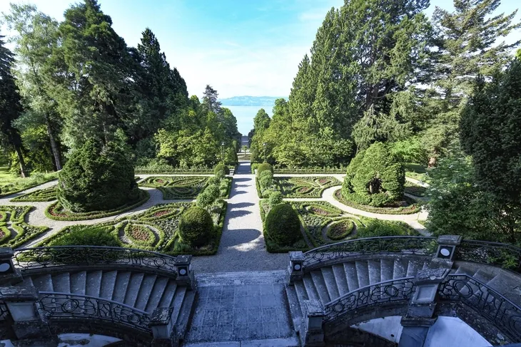 Blick von der Treppe der Villa Alwind, in der Ferienanlage Lindau, auf einen symmetrisch angelegten historischen Garten mit Hecken und Wegen vor Bäumen. Dahinter sieht man den Bodensee unter leichtbewölktem blauen Himmel.