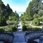 Blick von der Treppe der Villa Alwind, in der Ferienanlage Lindau, auf einen symmetrisch angelegten historischen Garten mit Hecken und Wegen vor Bäumen. Dahinter sieht man den Bodensee unter leichtbewölktem blauen Himmel.