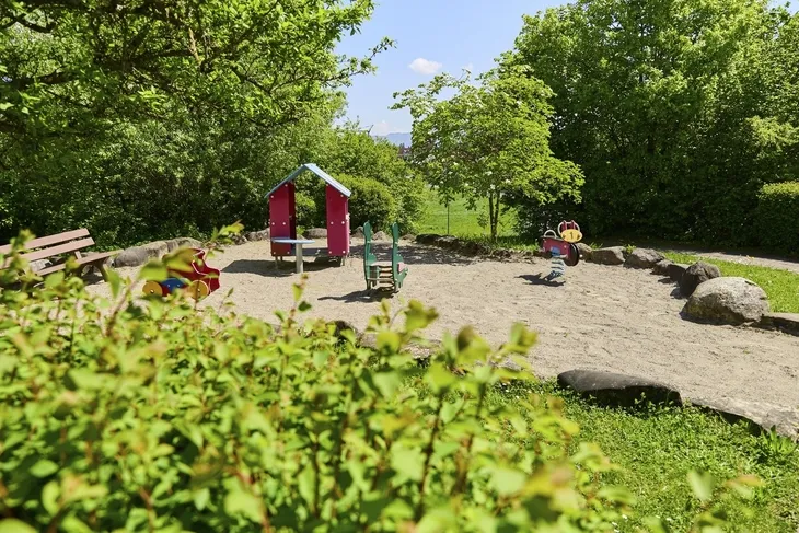 Kleiner Spielplatz mit Wiipptieren und Häuschen sowie einer Parkbank umgeben von Bäumen bei sonnigem Wetter in der Ferienanlage Lindau.