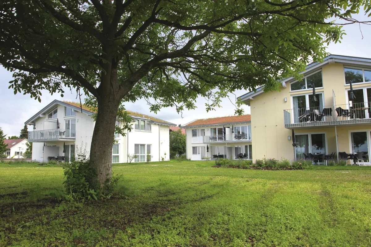 Drei Appartementhäuser an der Obstwiese, der Ferienanlage Lindau, mit Balkonen und Terrassen mit Outdoormöbeln ausgestattet und einem Baum auf einer grünen Wiese im Vordergrund.