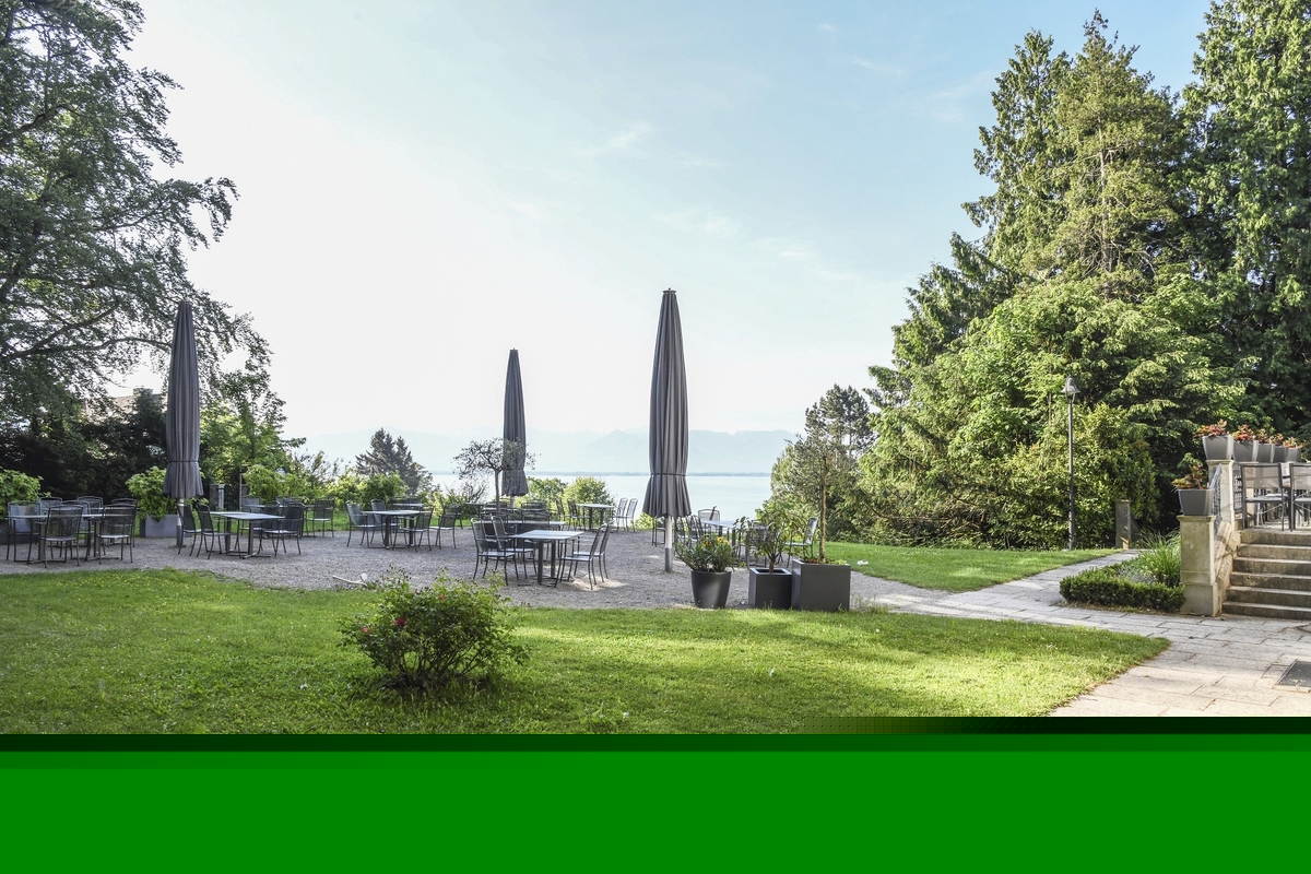 Biergarten der Villa Alwind, in der Ferienanlage Lindau, mit Tischen und Stühlen, sowie geschlossenen Sonnenschirmen, umgeben von Bäumen und einer Wiese mit Blick auf den Bodensee. bei leicht bewölktem Himmel.