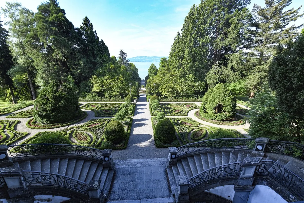 Blick von der Treppe der Villa Alwind, in der Ferienanlage Lindau, auf einen symmetrisch angelegten historischen Garten mit Hecken und Wegen vor Bäumen. Dahinter sieht man den Bodensee unter leichtbewölktem blauen Himmel.