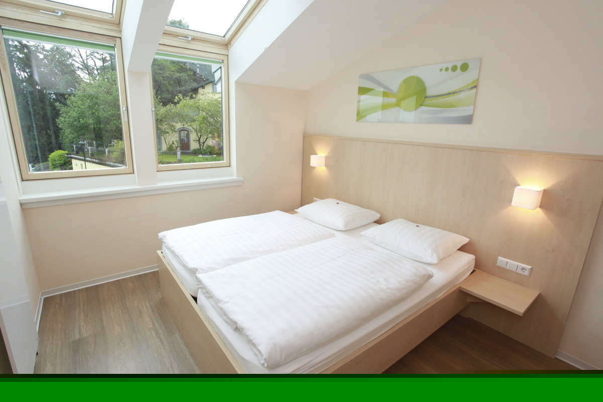 Helles Schlafzimmer in der Ferienanlage Lindau, im Haus an der Obstwiese, mit Doppelbett mit Lampen, senkrechten Fenstern und Dachfenstern, sowie einem Bild über dem Bett. .