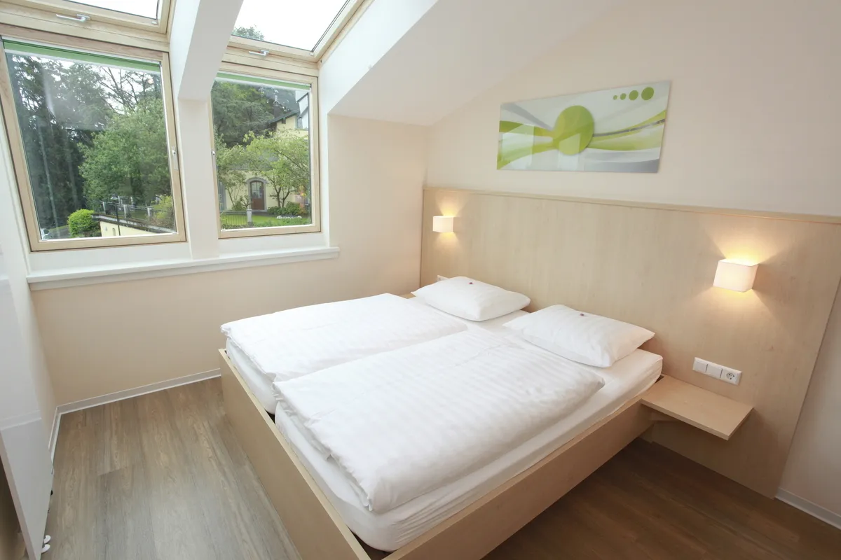 Helles Schlafzimmer in der Ferienanlage Lindau, im Haus an der Obstwiese, mit Doppelbett mit Lampen, senkrechten Fenstern und Dachfenstern, sowie einem Bild über dem Bett. .