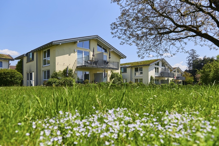 Zwei hellgelbe Appartementhäuser an der Obstwiese, der Ferienanlage Lindau, auf einer grünen und blühenden Wiese und einem Baum im Vordergrund.