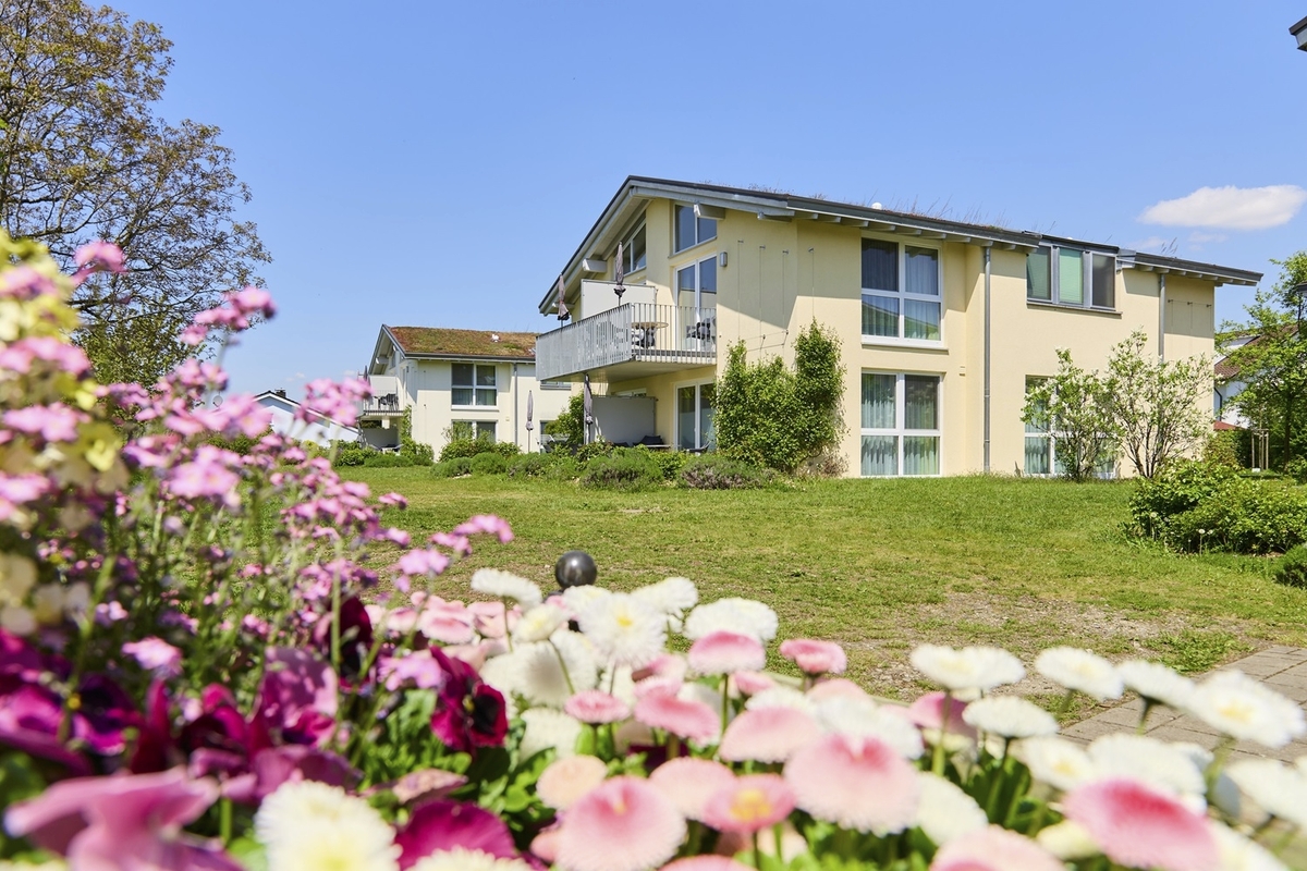 Zwei hellgelbe Appartementhäuser an der Obstwiese, der Ferienanlage Lindau, mit großem Balkon, auf einer Wiese mit rosa und lila blühenden Blumen im Vordergrund bei strahlend blauem Himmel.