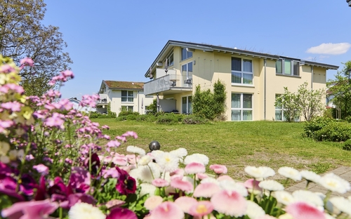 Zwei hellgelbe Appartementhäuser an der Obstwiese, der Ferienanlage Lindau, mit großem Balkon, auf einer Wiese mit rosa und lila blühenden Blumen im Vordergrund bei strahlend blauem Himmel.