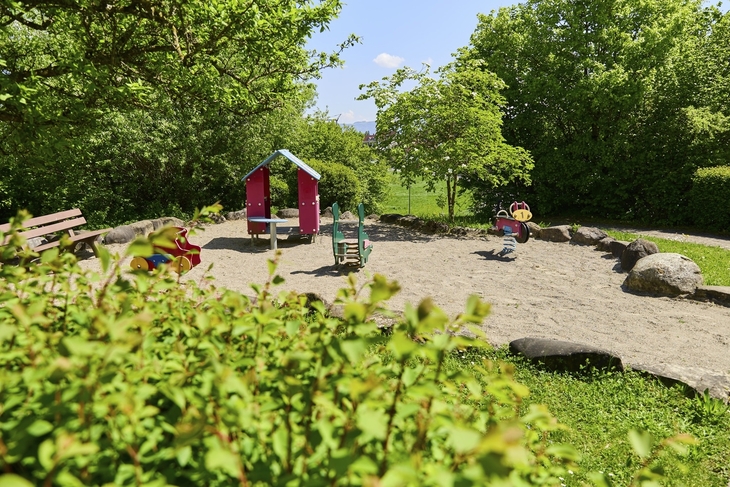 Kleiner Spielplatz mit Wipptieren und Häuschen sowie einer Parkbank umgeben von Bäumen bei sonnigem Wetter in der Ferienanlage Lindau.