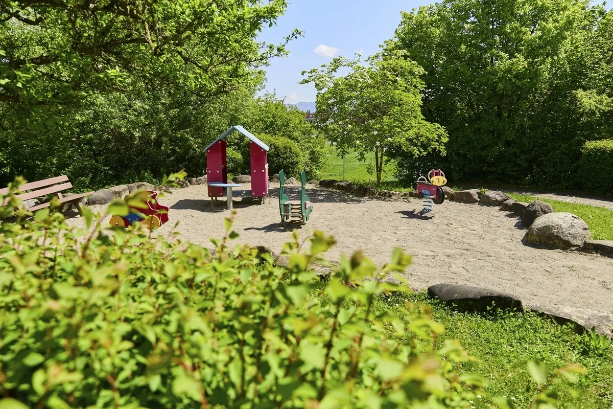 Kleiner Spielplatz mit Wipptieren und Häuschen sowie einer Parkbank umgeben von Bäumen bei sonnigem Wetter in der Ferienanlage Lindau.