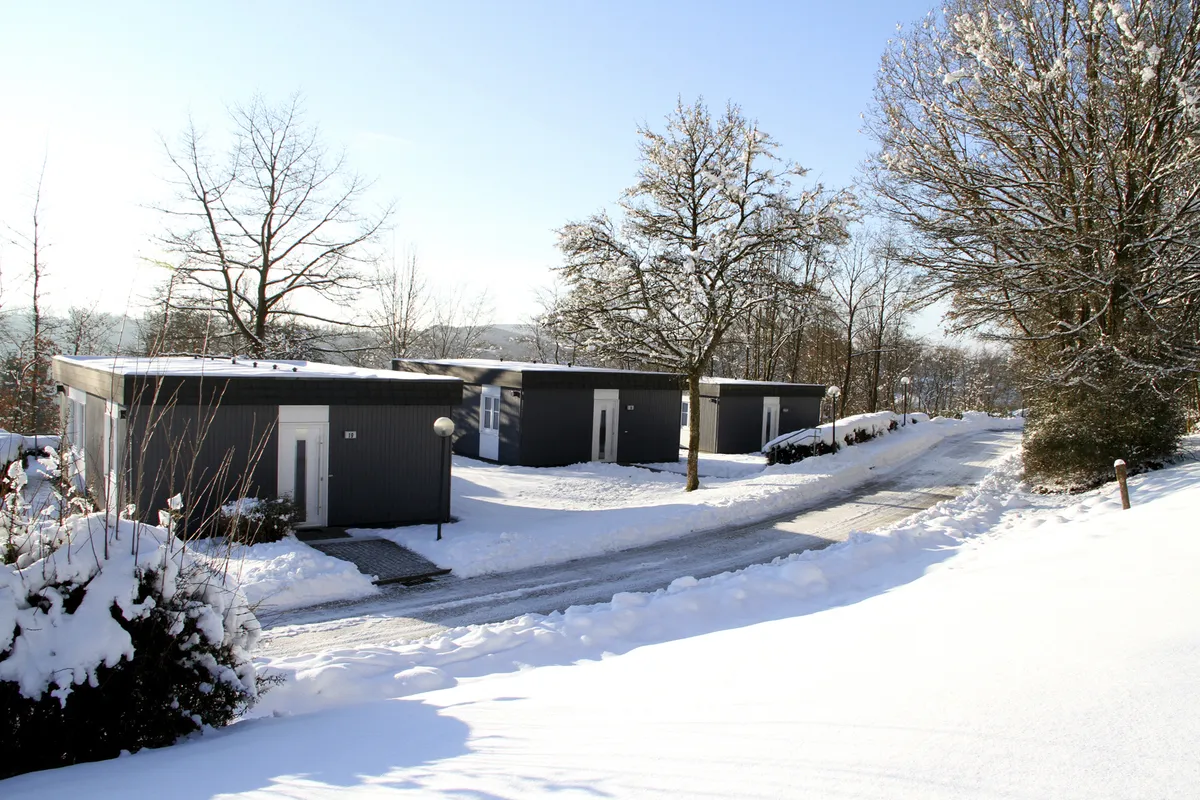 Bungalows der Ferienanlage Prüm in schneebedeckter Winterlandschaft mit Bäumen unter klarem Himmel.