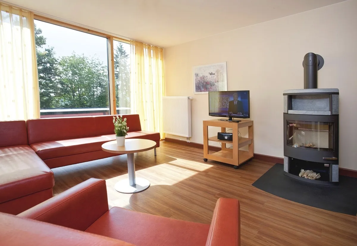 Wohnzimmer in der Ferienanlage Prüm, mit rotem Sofa und Sessel, kleiner Tisch, Holzofen, Fernseher und Blick auf Bäume durch große Fenster.