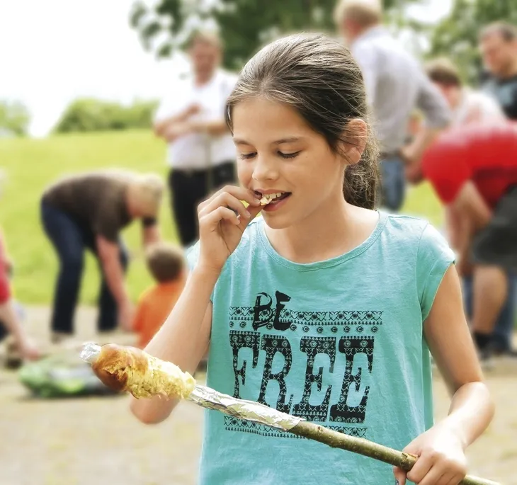 Mädchen isst Stockbrot auf einer Wiese in der Ferienanlage Prüm, Menschen im Hintergrund.