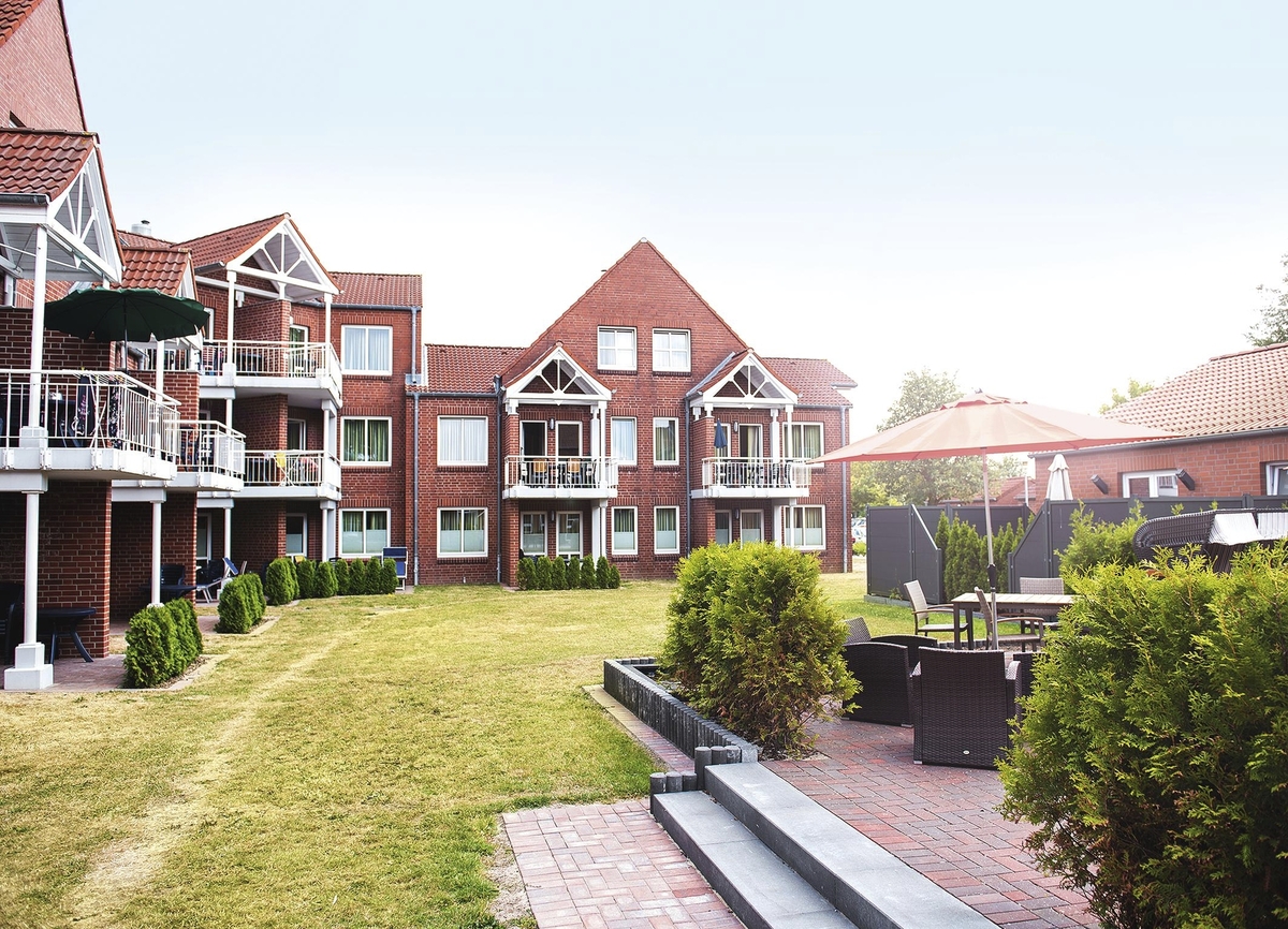 Außenaufnahme der Ferienanlage Büsum, bestehend aus aneinandergebauten roten Klinkergebäude mit spitzen Dächern und weißen Balkonen, vor sonnigem Himmel. Rechts im Bild eine Terrasse mit Sitzmöbeln und zwischen den Häusern und der Terrasse ein breiter Grünstreifen. 