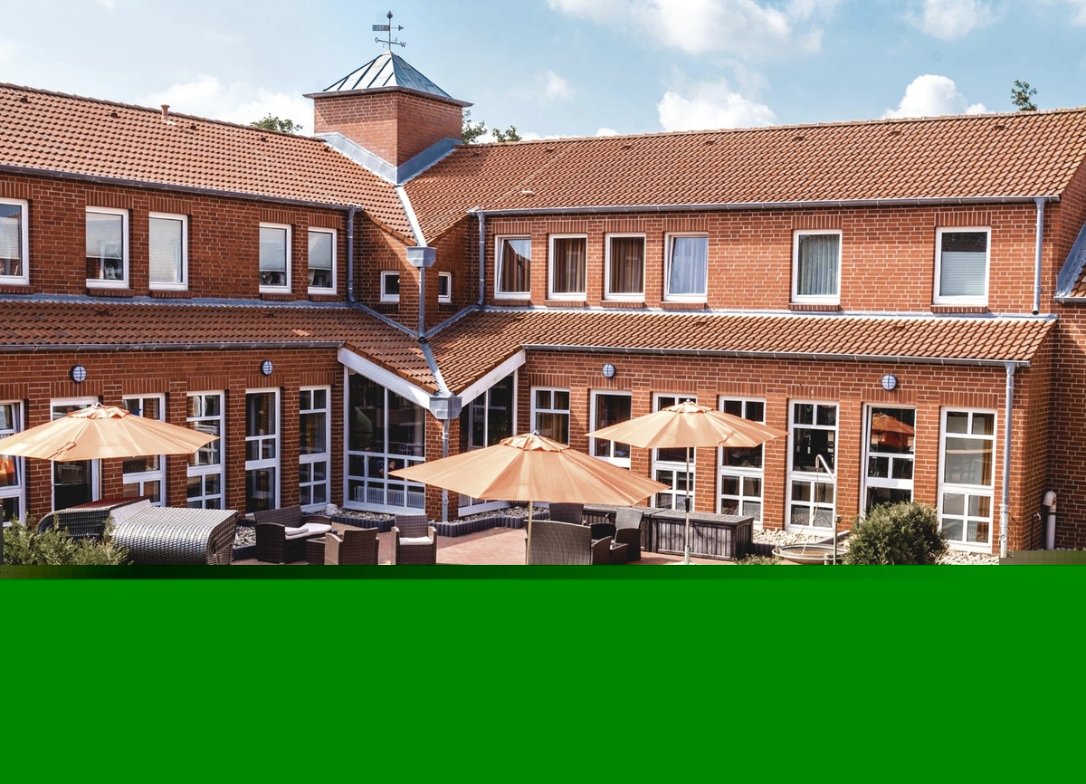 Rotes rechtwinkliges Backsteingebäude der Ferienanlage Büsum mit Terrasse, Sitzmöbeln und Sonnenschirmen bei sonnigem Wetter.