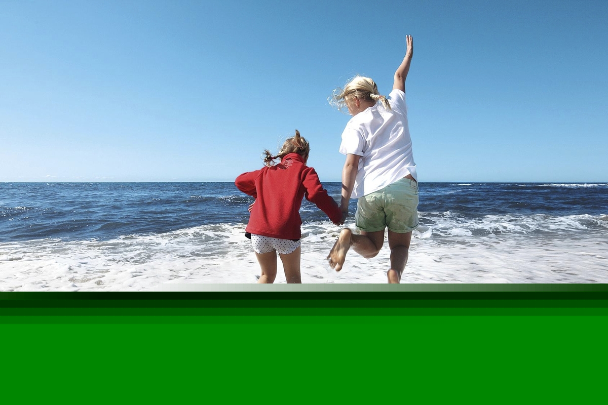 Zwei Kinder, in kurzer Hose und T-Shirt, springen vergnügt Hand in Hand in das schäumende Meer vor blauem Himmel.