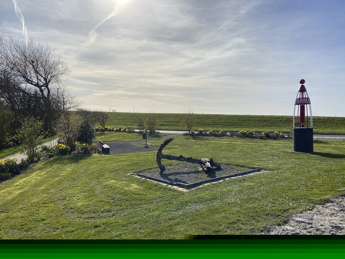 Blick auf eine Boje und Anker am Deich in Harlesiel, umgeben von Rasen und Bäumen.