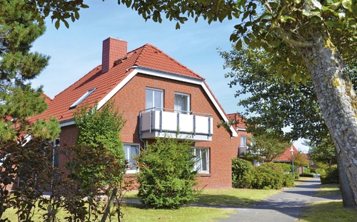 Rotes Ferienhaus mit Garten und Balkon in Ferienanlage Harlesiel, umgeben von grünen Bäumen und einem Gehweg.