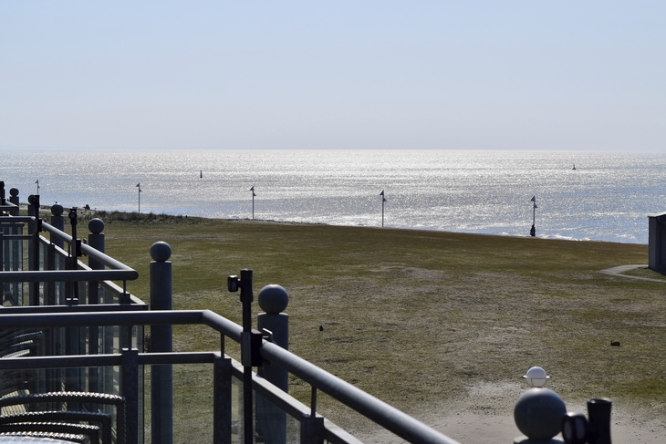 Blick vom Balkon der Ferienanlage Norderney auf das sonnige Meer mit hellblauem Himmel und grasbewachsenen Uferbereich.