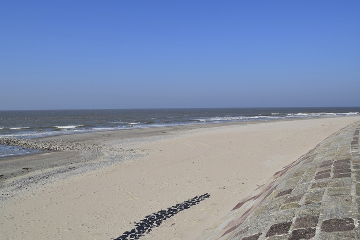 Ansicht eines weiten Sandstrands, rechts durch Steine befestigt und links das Meer, bei strahlend blauem Himmel.