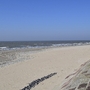 Ansicht eines weiten Sandstrands, rechts durch Steine befestigt und links das Meer, bei strahlend blauem Himmel.