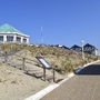 Befestigter Weg vom Strand zur Ferienanlage Norderney vor blauem Himmel. Links im Bild führen Treppenstufen über Dünen zu einem achteckigen weißen Pavillon mit grünem Dach. 