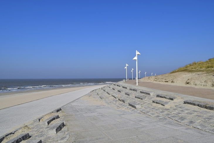 Strandpromenade mit Laternen und befestigtem Weg, mit Blick auf Meer und Strand, bei strahlend blauem Himmel.