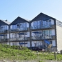 Dreistöckige aneinander gebaute Häuser mit spitzen Dächern, der Ferienanlage Norderney, mit Balkonen, ausgestattet mit Sitzmöbeln und blauen Sonnenschirmen vor klarem Himmel und grünem Bewuchs im Vordergrund.