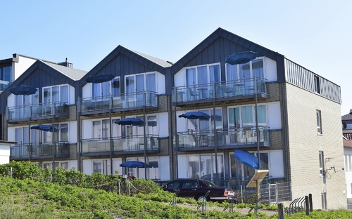 Dreistöckige aneinander gebaute Häuser mit spitzen Dächern, der Ferienanlage Norderney, mit Balkonen, ausgestattet mit Sitzmöbeln und blauen Sonnenschirmen vor klarem Himmel und grünem Bewuchs im Vordergrund.