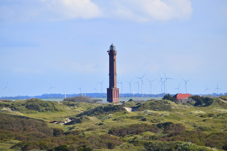 Leuchtturm auf Norderney mit Windrädern im Hintergrund und grüner, leicht hügeliger Landschaft im Vordergrund.