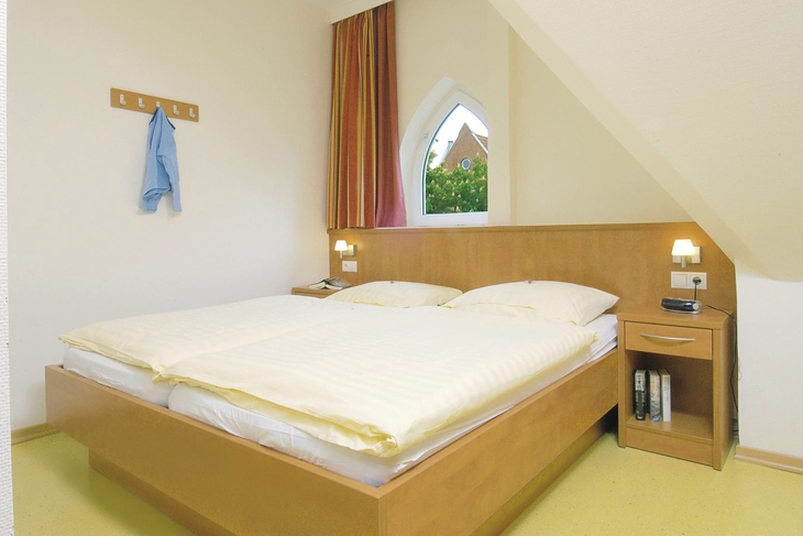 Schlafzimmer mit Doppelbett, Nachttischen und Nachttischlampen, einer Hakenleiste an der Wand, sowie einem spitz zulaufenden Fenster mit einem bunten Vorhang, in der Ferienanlage in Wyk auf Föhr.