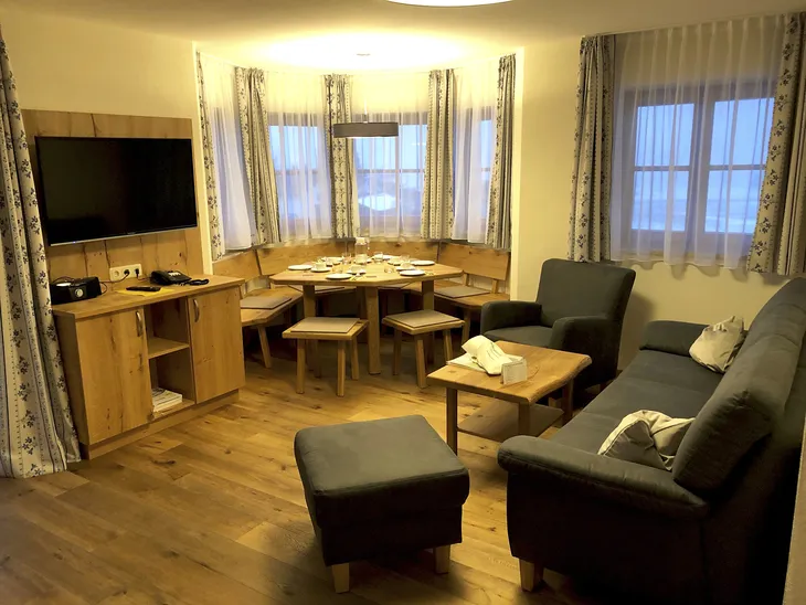 Wohnzimmer mit Sofa, Fernseher, Esstisch und Stühlen, Fenster mit Vorhängen.