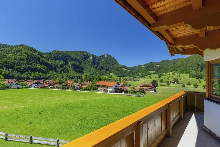 Blick vom Balkon auf eine grüne Wiese und Berge bei sonnigem Wetter.