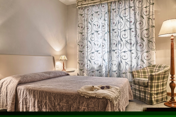 Schlafzimmer mit Doppelbett, gefliester Boden, Sessel und Vorhänge mit floralem Muster.