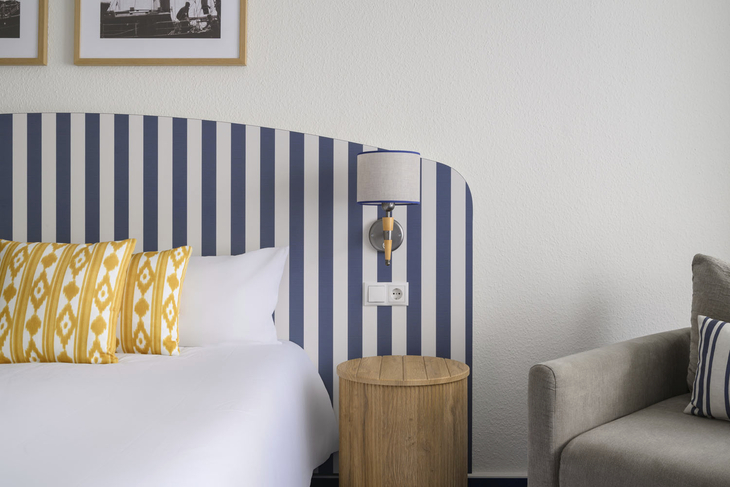 Hotelzimmer mit gestreiftem Kopfteil, gelben Kissen und Tischlampe.