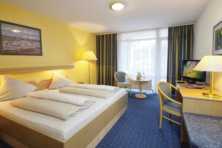 Hotelzimmer mit Bett, Schreibtisch, Stuhl und Fernseher, gelbe Wände und blauem Teppich.