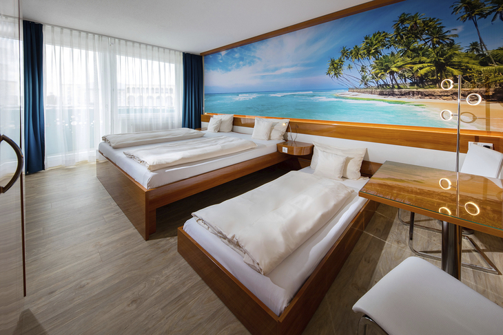 Hotelzimmer mit drei Betten, Meeresmotiv an der Wand, helle und moderne Einrichtung.