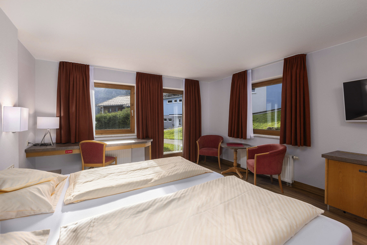 Doppelzimmer mit zwei Betten, Tisch, Stühlen und Fenstern mit Vorhängen.