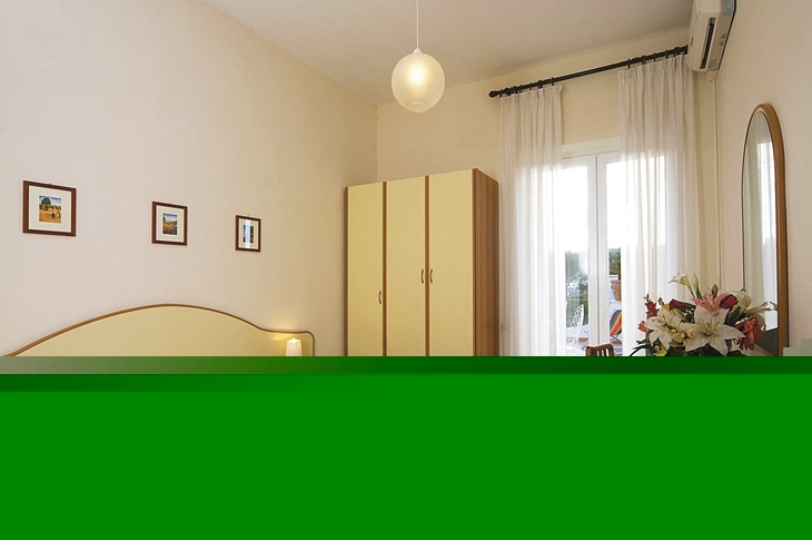 Helles Hotelzimmer mit Bett, Schrank, Tisch, Spiegel und Blumen.