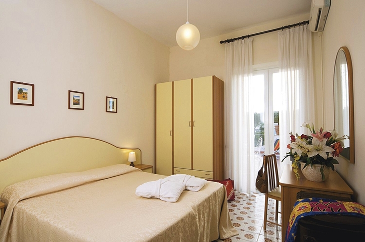 Helles Hotelzimmer mit Bett, Schrank, Tisch, Spiegel und Blumen.