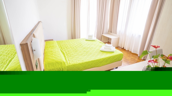Helles Hotelzimmer mit grünem Bett, Pflanzen und Fenstervorhängen.