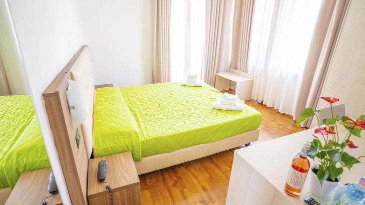 Helles Hotelzimmer mit grünem Bett, Pflanzen und Fenstervorhängen.