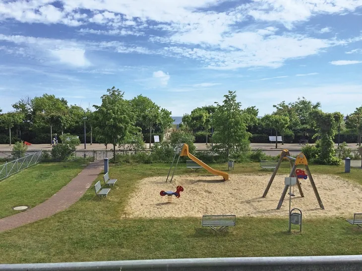 Spielplatz mit Rutsche und Wippe, umgeben von Bäumen und Wiese.