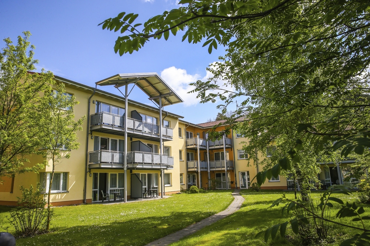 Dreistöckiges Wohngebäude mit Balkonen in einer grünen Umgebung bei Sonnenschein.