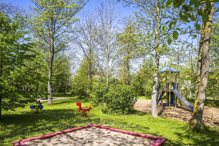 Spielplatz im Grünen mit Rutsche, Schaukelpferd und Bäumen im Sonnenschein.